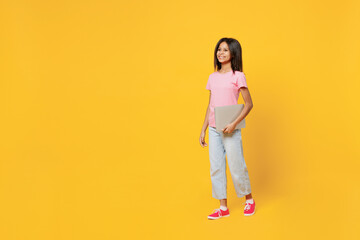 Fototapeta premium Full body happy little kid girl of African American ethnicity 12-13 years old in pink t-shirt hold closed laptop pc computer walk go isolated on plain yellow background. Childhood lifestyle concept.