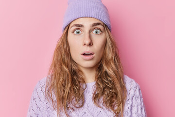 Shocked startled Caucasian woman has eyes popped out looks with amazement holds breath wears purple hat and knitted jumper isolated over pink background. Human reactions and emotions concept