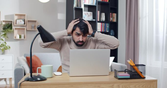 Young Arab tired and sad man working at laptop and getting bad news or result in job. At home. Arabian upset male freelancer at computer. Gamer loosing in game online. Lockdown concept. Lost money.