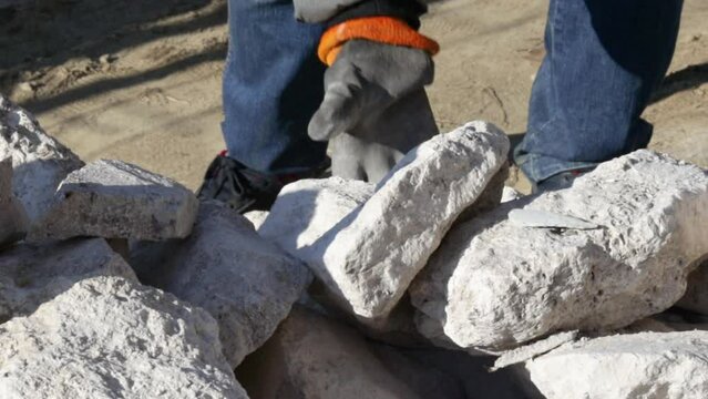 stack of rubble stones outside, sunny day, and a man is taking some pieces. young man, constructor,master builder, takes the decorative pieces of stone with hands in gloves. white ragstone rubble 