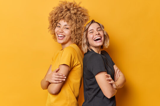 Positive Two Young Pretty Women Stand Back To Each Other Keep Arms Folded Laugh Happily Dressed In Casual T Shirts Laugh At Something Funny Isolated Over Yellow Background. Friendship Concept