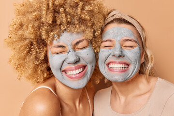 Portrait of overjoyed women smile broadly keep eyes closed show white teeth apply moisturising clay mask on face undergo skin care procedures isolated over brown background express positive emotions