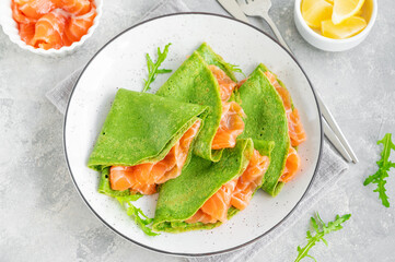 Green spinach crepes or pancakes with smoked salmon and soft cheese on a plate on the gray background. Copy space.