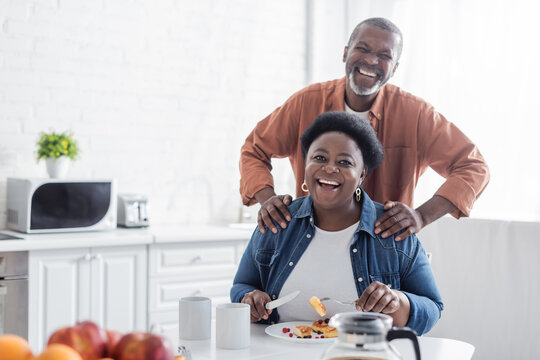 happy and senior african american man standing behind wife during breakfast.