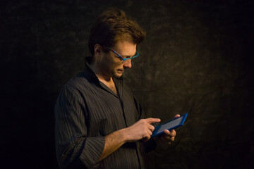 Retrato de hombre joven con camisa oscura, anteojos azules y tablet azul sobre fondo negro....