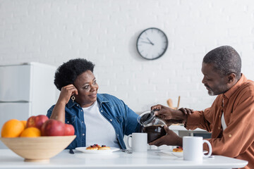 senior african american man pouring coffee to happy wife during breakfast.