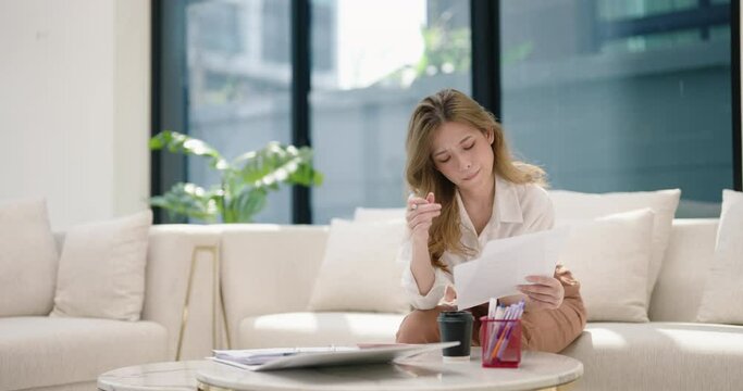 Young Asian woman sitting on couch holds paper receipt calculates monthly expenses using calculator check utility bills managing family budget, accountant do paperwork job at home concept.