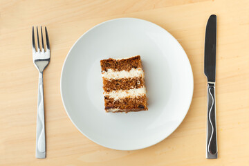 A piece of carrot sponge cake on white plate on wooden table with a metal cutlery fork and knife. background for restaurant menu with copy space for your text