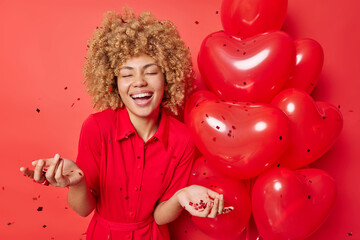 Glad young woman with curly bushy hair wears dress stands near heart shaped inflated balloons against red background with confetti laughs happily enjoys festive event awaits for party to start
