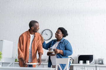 happy african american man holding pancakes on plate and looking at wife with coffee pot.