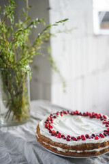 A delicious carrot cake stands on the table, garnished with white mascarpone cream and cranberries. A beautiful dessert for every day. There are greens in a vase on the table. Spring still life