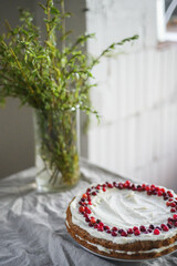 A delicious carrot cake stands on the table, garnished with white mascarpone cream and cranberries. A beautiful dessert for every day. There are greens in a vase on the table. Spring still life