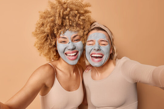 Overjoyed women laugh gladfully pose next to each other have fun make selfie photo keep arms outstreched take care of skin wears nourishing clay beauty masks isolated over brown wall. Beauty concept