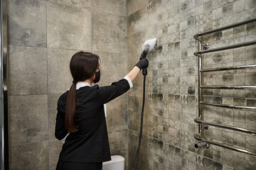 Cleaning service company employee in rubber gloves cleaning tiles in bathroom with professional steam cleaner equipment