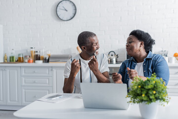 senior african american husband and wife rejoicing and looking at each other near laptop.