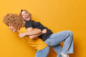 Energetic young women lean at backs of each other give piggy back ride dressed in casual t shirt and jeans feel overjoyed isolated over yellow backgrounf have fun together have friendly relations