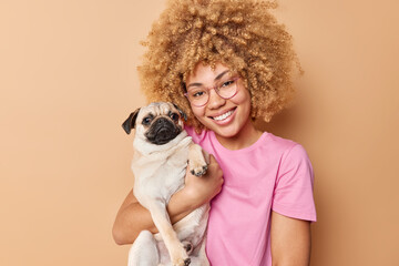 Portrait of cheerful young woman hugs her favorite pug dog smiles gladfully expresses love and care...