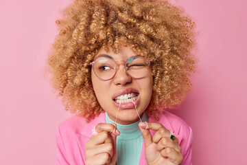 Dental health care concept. Beautiful young woman with curly bushy hair cleans teeth with dental floss wears transparent eyesglasses isolated over pink background shows how to floss correctly - obrazy, fototapety, plakaty