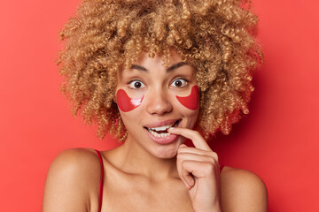 Surprised curly haired young woman bites finger looks excited at camera applies hydrogel pads under eyes for rejuvenation and skin moisturising stands bare shoulders against vivid red background