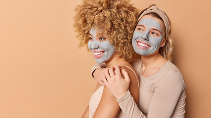 Cheerful friendly women embrace and express care to each other apply beauty clay mask on face for rejuvenation and reducing blackheads stand sideways isolated over beige background mock up space