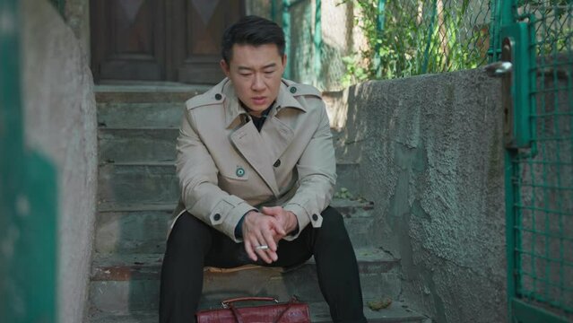 Stressful young asian businessman adult sitting outside house on stairs smoking cigarette and overthinking. Frustrated man.
