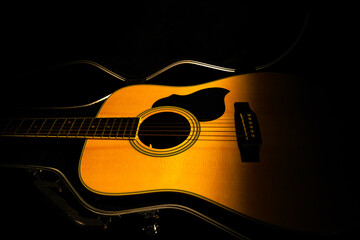 Yellow acoustic guitar lying in a hard case in the dark on a black background. Wooden stringed instrument illuminated by a beam of light. Guitar in cover close up. Rock, folklore, country music. - obrazy, fototapety, plakaty