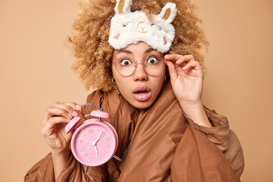 Shocked curly haired woman wakes up after seeing nightmare stares bugged eyes overslept work holds alarmclock keeps hand on rim of spectacles wears sleepmask on forehead wrapped in soft blanket