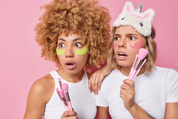 Shocked scared women stare away apply hydrogel patches under eyes pose with cosmetic brushes undergo beauty procedures dressed in casual white t shirts isolated over pink background. Cosmetology