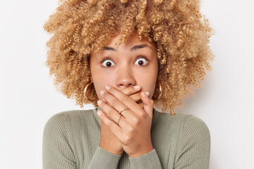 Shocked emotional young woman with curly hair covers mouth with hands tries to be speechless reacts on horrible scene wears casual turtleneck isolated over white background. Unbelievable news