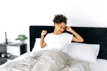 Obraz na płótnie Canvas Morning awakening. Happy lovely african american curly-haired girl in a white t-shirt, wakes up, does sipping her hands, starts a new day with a great mood, smiles, basking in bed