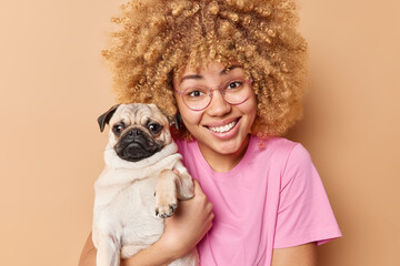 Portrait of happy European woman with blonde curly hair poses with pug dog being glad pet owner...