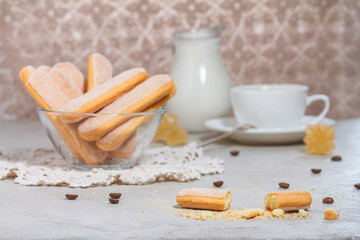 Fototapeta na wymiar Traditional italian savoiardi or ladyfingers biscuits in a glass bowl and pieces of cookies and sugar on the table, a white cup of coffee with a spoon and a jug of milk in the background