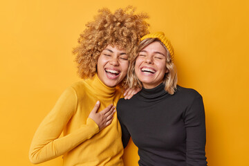 Sincere human emotions concept. Positive overjoyed young women have fun laugh gladfully smile toothily cannot stop laughing stand closely to each other dressed casually isolated over yellow wall