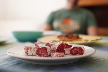 Diced Typical Spanish Fuet Cubes on a White Plate at a Friends Meal with out-of-focus Cheese and Human Figure in the Background