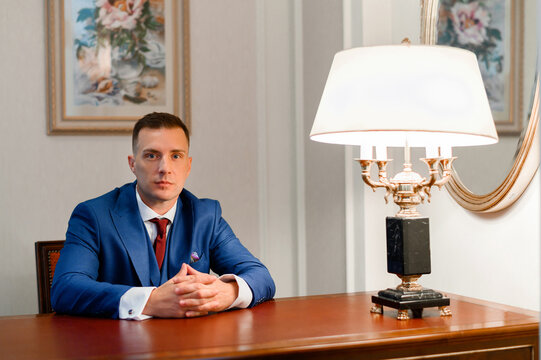 Front view of elegant businessman in trendy suit, keeping hands together, seriously looking at camera while sitting at wooden table with vintage lamp on background of wall with picture