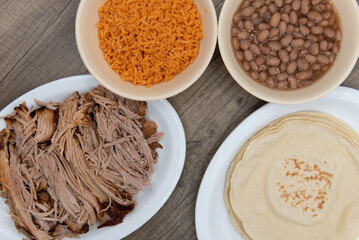 Overhead view of hearty Mexican meal spread out with carnitas pork, tortillas, rice and beans to...