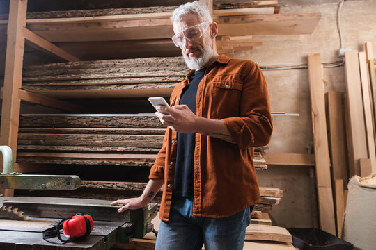 bearded furniture designer using smartphone near workbench in workshop.