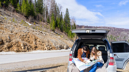 A young couple argues where to go on a trip sitting at the trunk of a car parked by the side of the road and reading a road map against the backdrop of mountains and roads. High quality photo