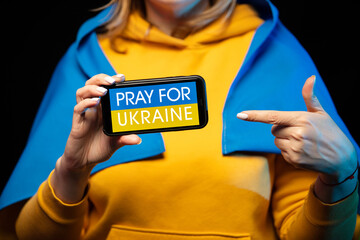 Beautiful Ukrainian girl with a state flag and smartphone in hands with a call PRAY FOR UKRAINE....
