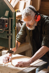 bearded woodworker in goggles cutting plank on band saw.