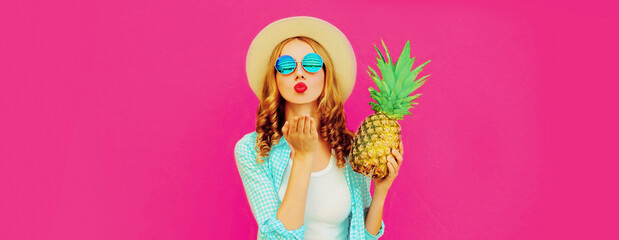 Fototapeta na wymiar Summer portrait of beautiful young woman with pineapple blowing her red lips sending sweet air kiss wearing straw hat, sunglasses on pink background