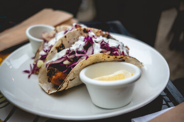 Closeup of white plate with gourmet 'tacos' (corn 'tortillas' with onion, fried chicken, sauces, vegetables), lemon and sauce in a table in the day