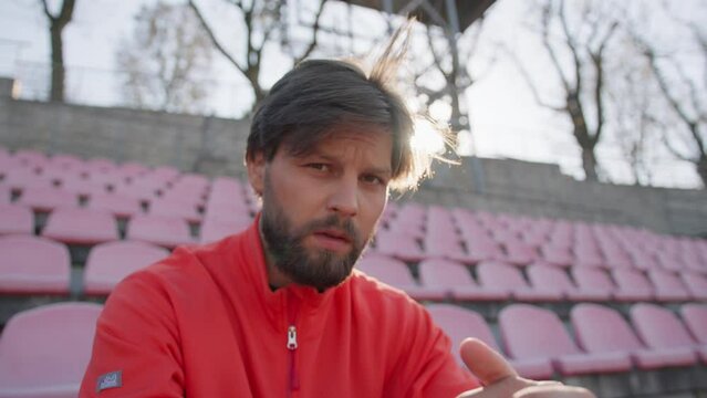 Outdoor portrait of attractive bearded guy wearing professional sport clothing sitting on tribune seats. Sportsman. Training people. Workout. Stadium.