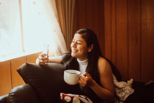 Mujer joven tomando caf&eacute; y mirando su tel&eacute;fono. Concepto de tecnolog&iacute;a y hogar. 