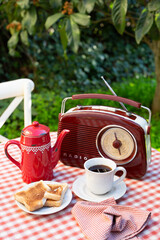 A white cup with coffee, red coffee pot and radio are on a table in a summer garden. Outdoor breakfast theme. A vertical image.