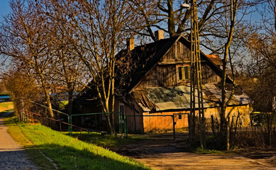 Ostrowiec - zabytkowy historyczny drewniany dom wśród drzew , wczesną wiosną , podświetlony zachodzącym słońcem . Chata z desek z dachem krytym papą i z dwoma kominami .