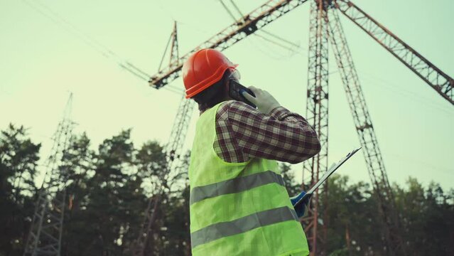 Electrical engineer is working. Talk on phone. Checking power grid. Energy. Industry. Electricity engineer beneath pylon talking on phone and checking data on tablet with documents of object
