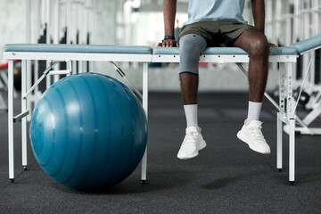 Low section shot of African American man sitting on massage bed in rehabilitation clinic - Powered by Adobe