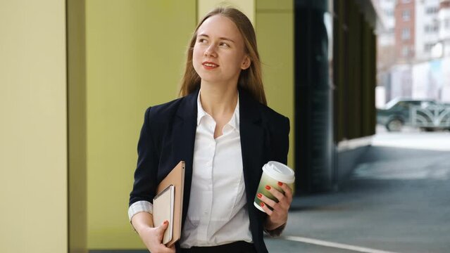 Confident young woman in suite and white shirt walking in city near office, holding laptop, papers and coffee cup. Student, startup successful businesswoman, junior manager of company.