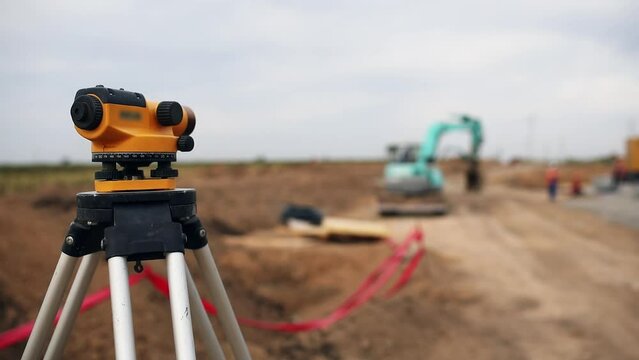 Optical level mounted on tripod against machinery operating at rural site. Excavator digs trench for laying power cables on cloudy day close view
